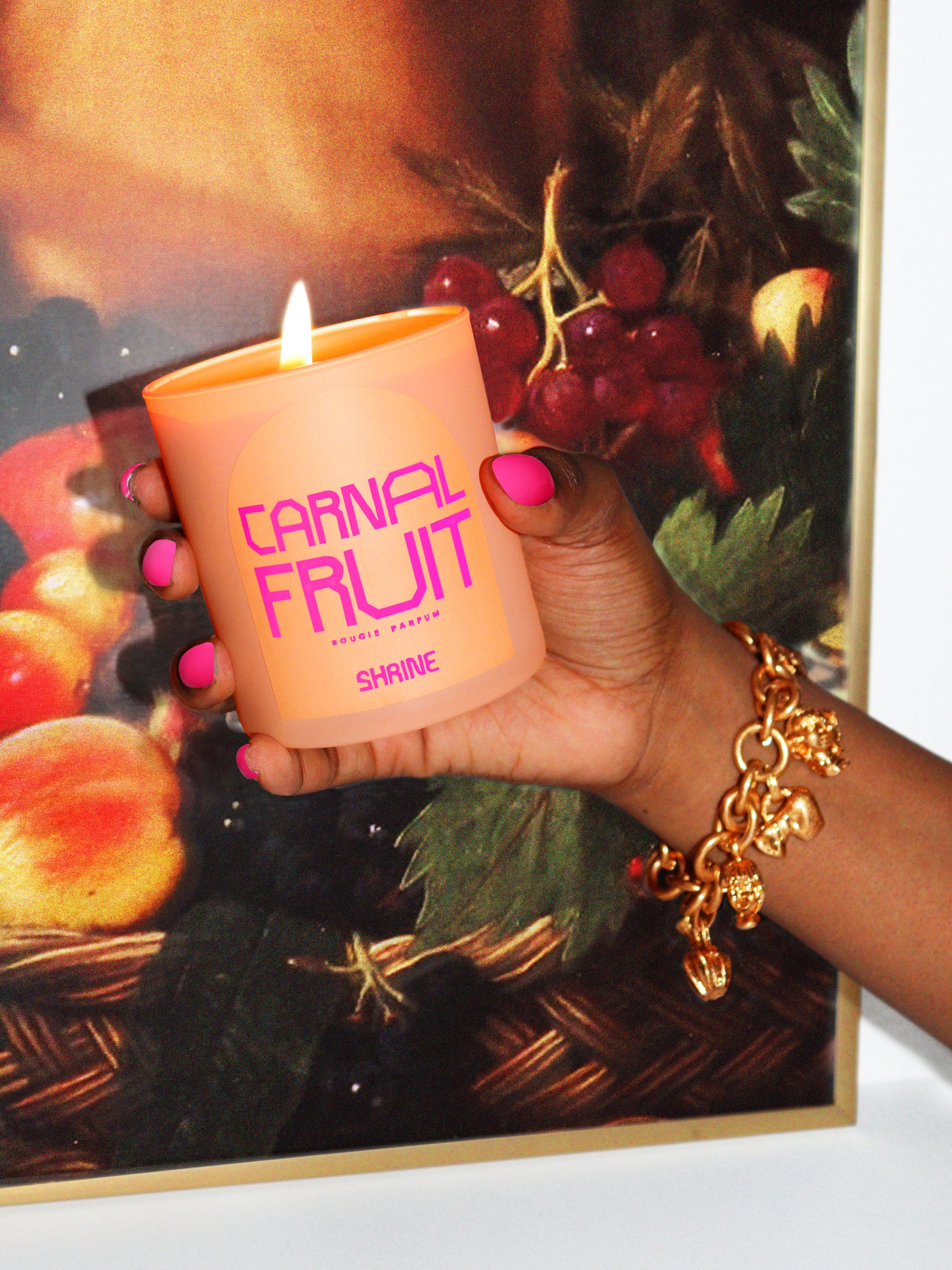 shrine carnal fruit monochromatic peach candle with neon pink text design in a hand with neon pink nails and gold charm bracelet in front of an oil painting of a fruit basket shop shrine